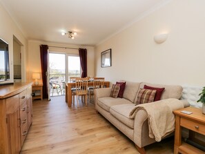 A cozy living room with a wooden floor, a beige sofa, a wooden dining table with chairs, and a TV on a wooden cabinet.