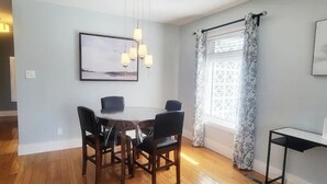 A dining area with a round table and four chairs, a hanging light fixture, a framed picture on the wall, and a window with patterned curtains.