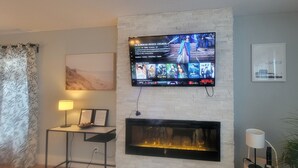 A modern living room with a flat-screen TV mounted on a white brick wall, a fireplace below, a side table with a lamp, and a large plant.