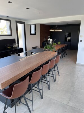 Une cuisine moderne dotée d’un grand îlot central, de meubles noirs et d’un coin repas avec une longue table en bois et des chaises marron.