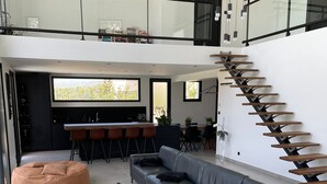Un salón moderno con un sofá gris, una mesa de centro de madera y un sillón de saco marrón. Se aprecia una escalera con escalones de madera y barandilla de vidrio.