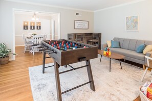 Uma sala de estar com mesa de futebol, um sofá cinza e um espaço de jantar com cadeiras e uma mesa.