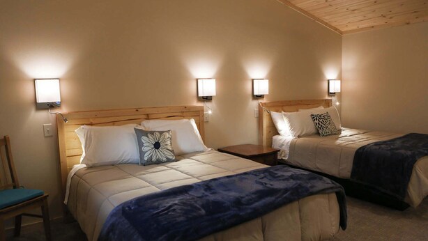 A hotel room with two beds, each with a headboard and bedside lamps. There is a chair and a small table in the room.