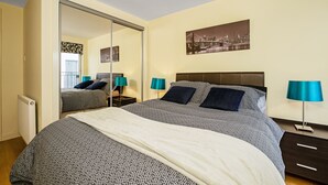 A bedroom with a bed, a nightstand, a lamp, a mirror, and a cityscape picture on the wall. - Bell Street Apartment (Glasgow)
