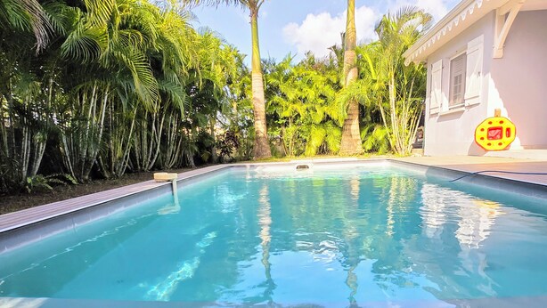 A swimming pool with a wooden deck, surrounded by lush greenery and a white building with windows.