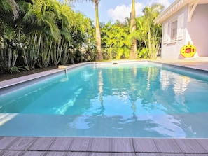 A swimming pool with a wooden deck, surrounded by lush greenery and a white building with windows. - Villa Carpe Diem - Swimming Pool - Sleeps 9 - 4 Bedrooms (Les Trois-Îlets)