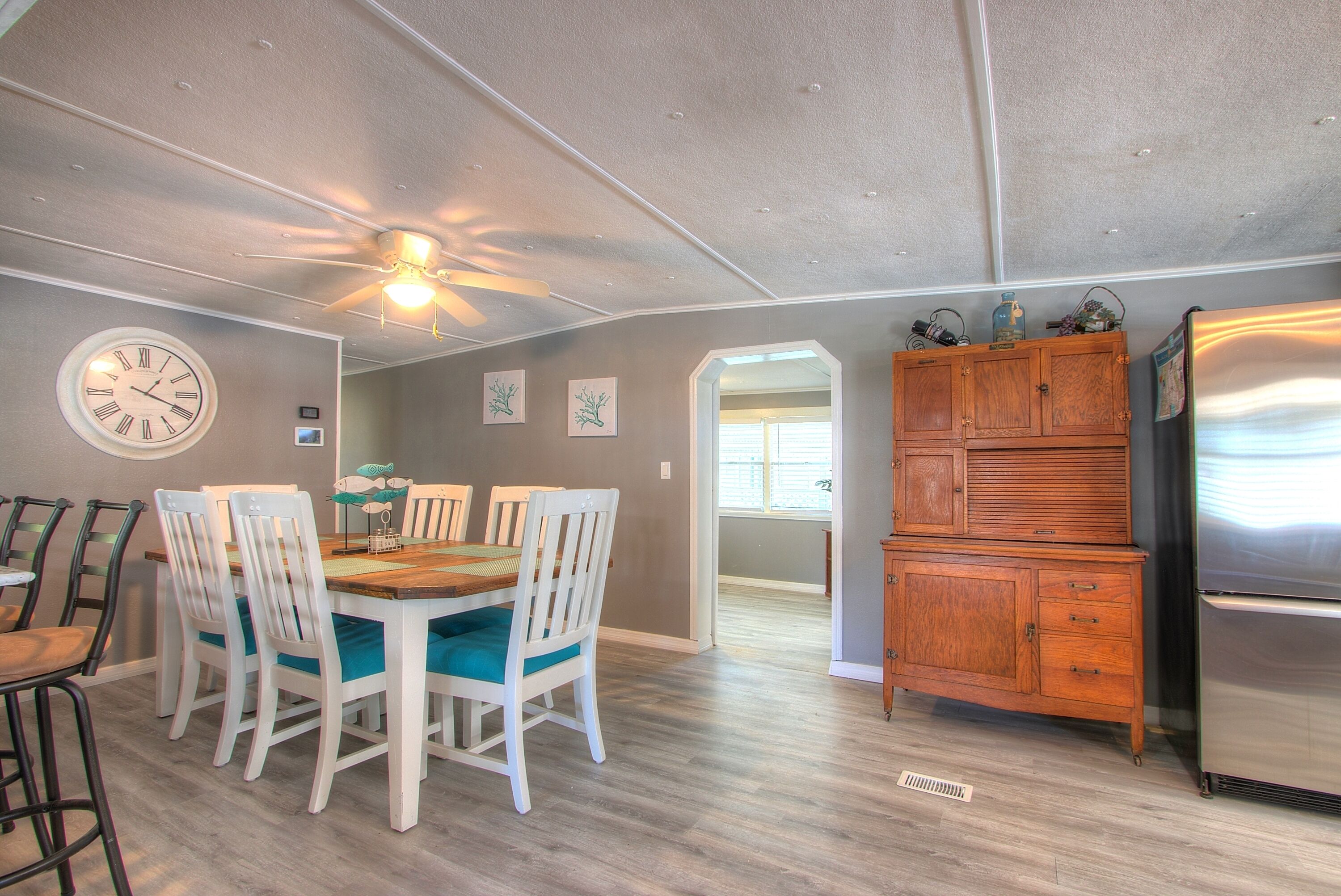 A dining area with a wooden table and white chairs, a clock on the wall, and a ceiling fan.