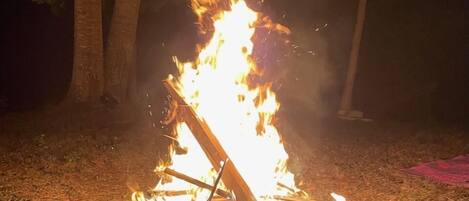 Una fogata con leña y ramas dispuestas en forma de tipi, rodeada de hierba y árboles.
