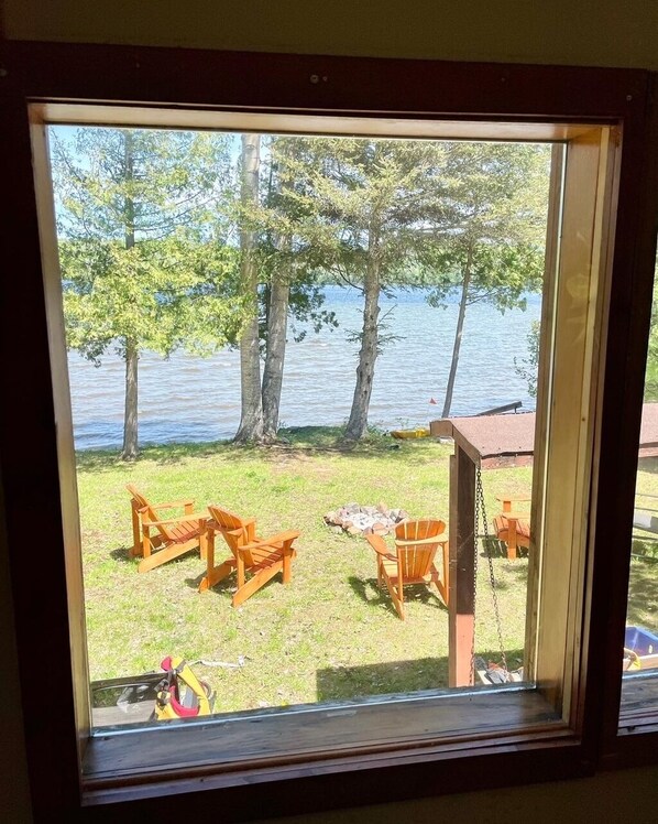 A window view of a lakeside outdoor seating area with wooden chairs and tables, a fire pit, and a bicycle. - Cozy, modern cottage lots of space, perfect waterfront for swimming / water fun! (Barry's Bay)