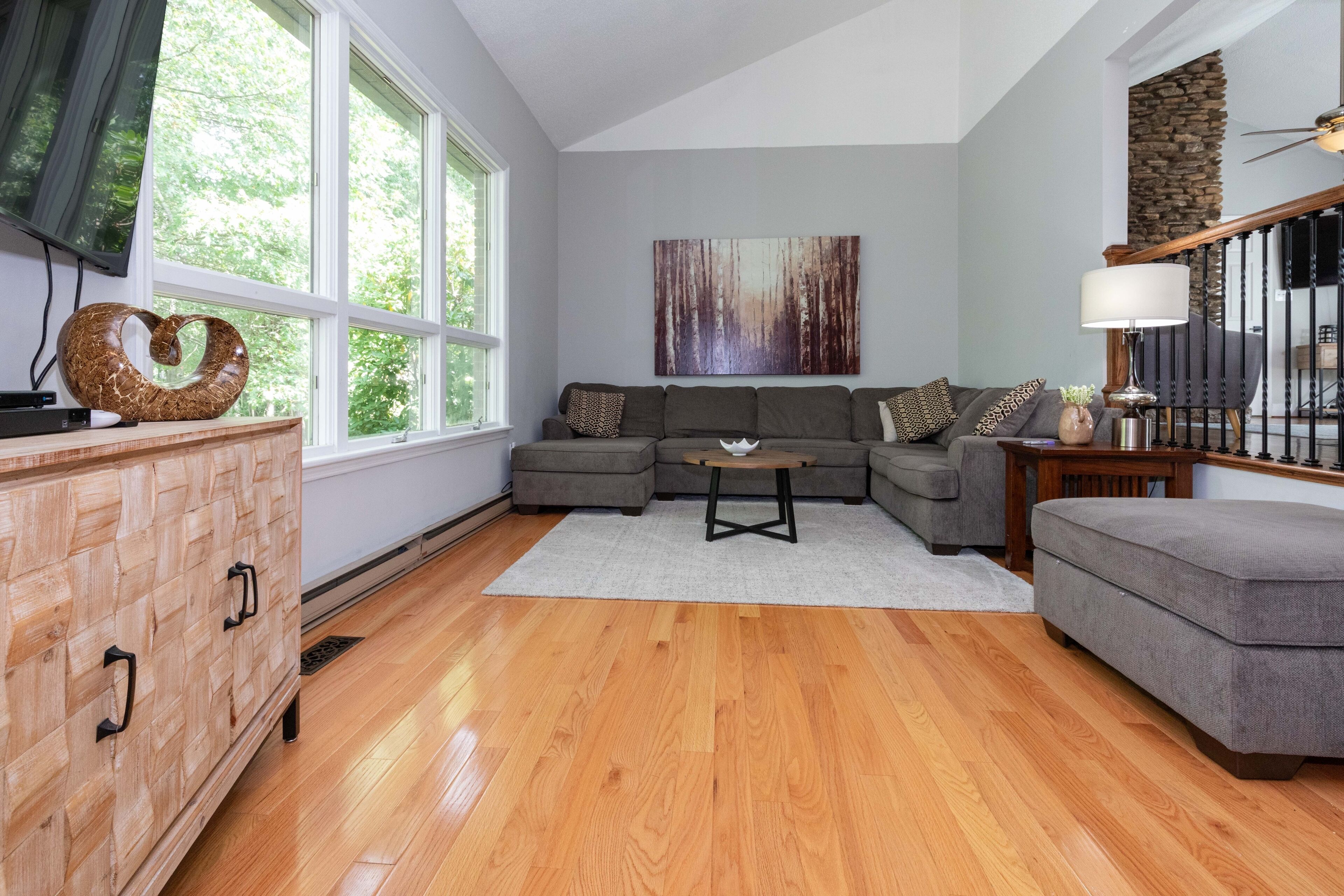A living room with a sectional sofa, a wooden cabinet, a coffee table, and a large window with a view of trees.