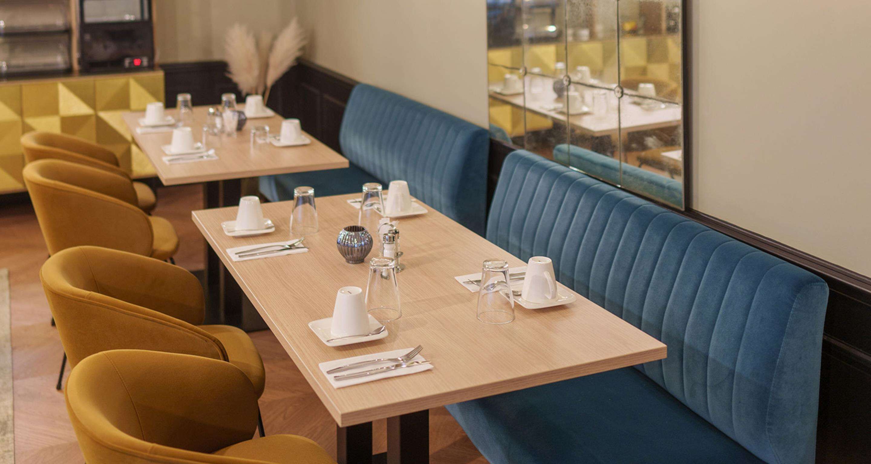 A restaurant with blue upholstered booths and yellow chairs, tables set with white plates, glasses, and cutlery.