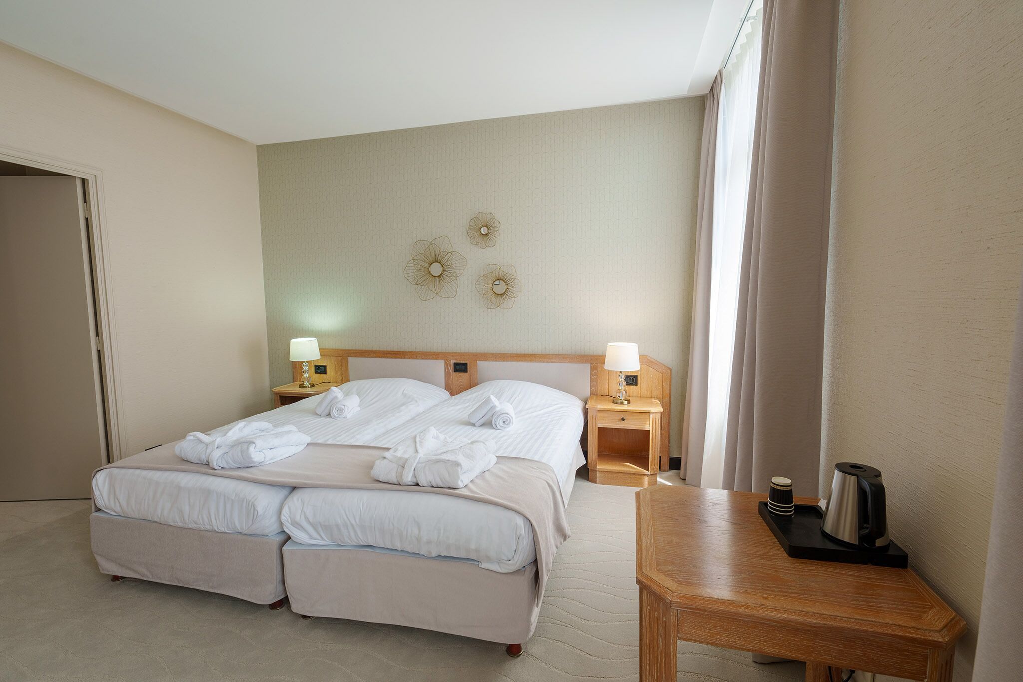 A hotel room with a double bed, two bedside tables with lamps, a wooden nightstand, a coffee maker, and a wall with three decorative wall art pieces.
