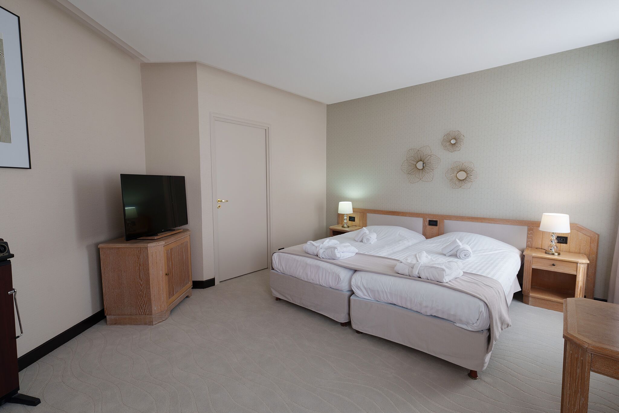 A hotel room with a bed, a TV on a wooden cabinet, a bedside table with a lamp, and a wall-mounted decorative piece.