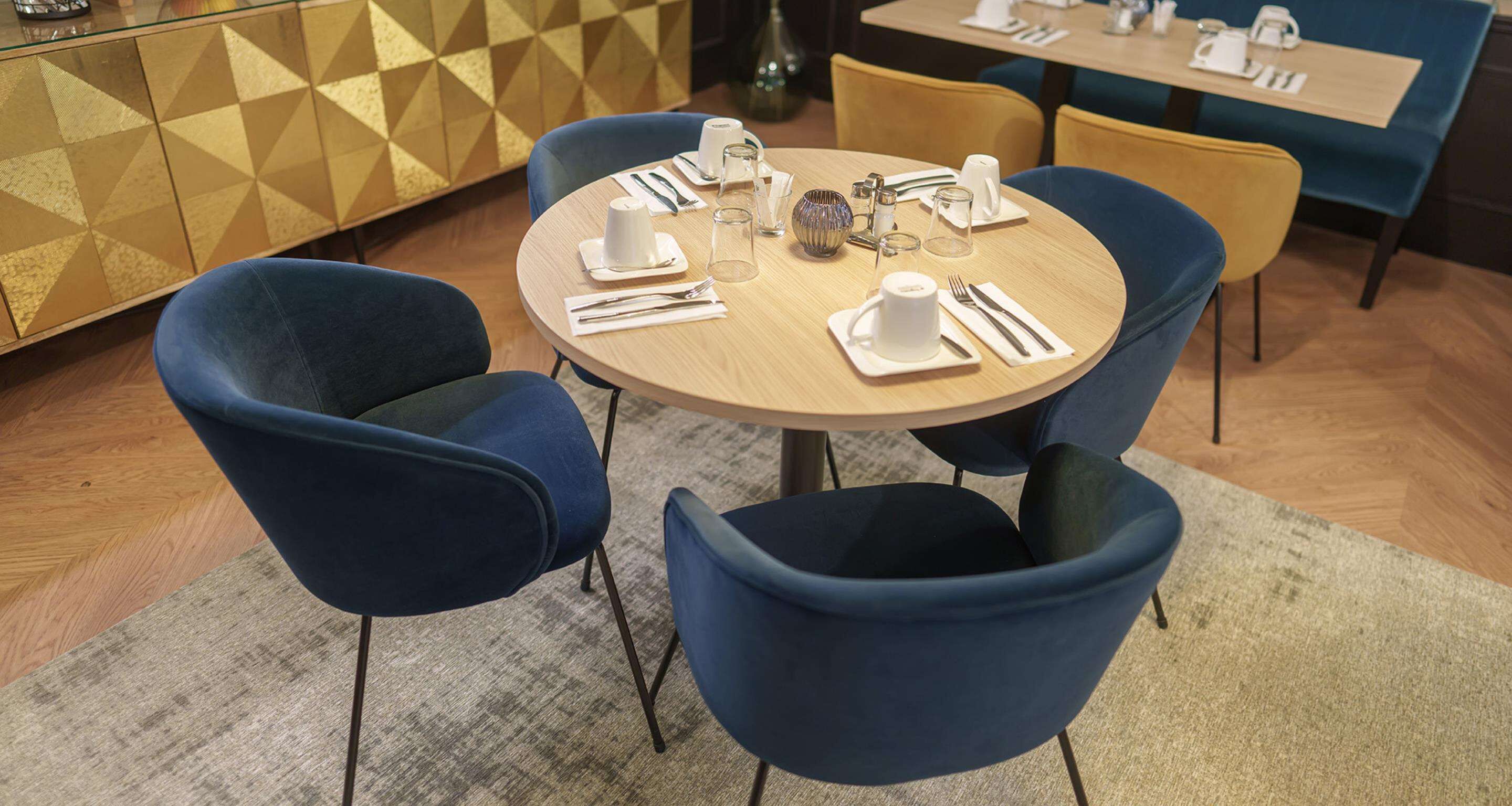 A round table set with white plates, cups, and cutlery, surrounded by blue chairs in a modern dining area with geometric patterned walls.