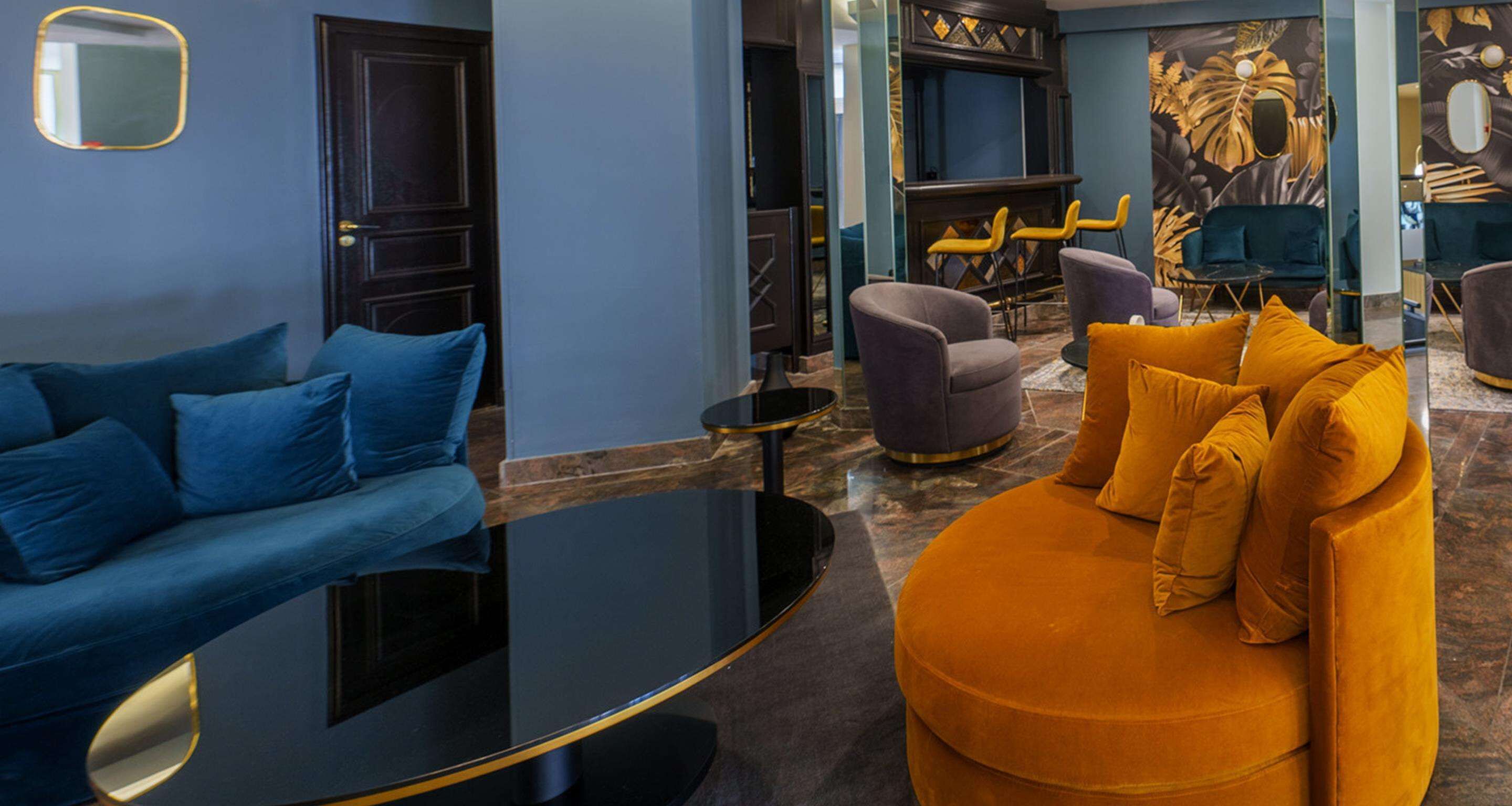 A modern living room with a blue sofa, a round orange ottoman, and a black coffee table. There are two armchairs and a small table with yellow chairs. The room has a dark wooden door and a mural on the wall.
