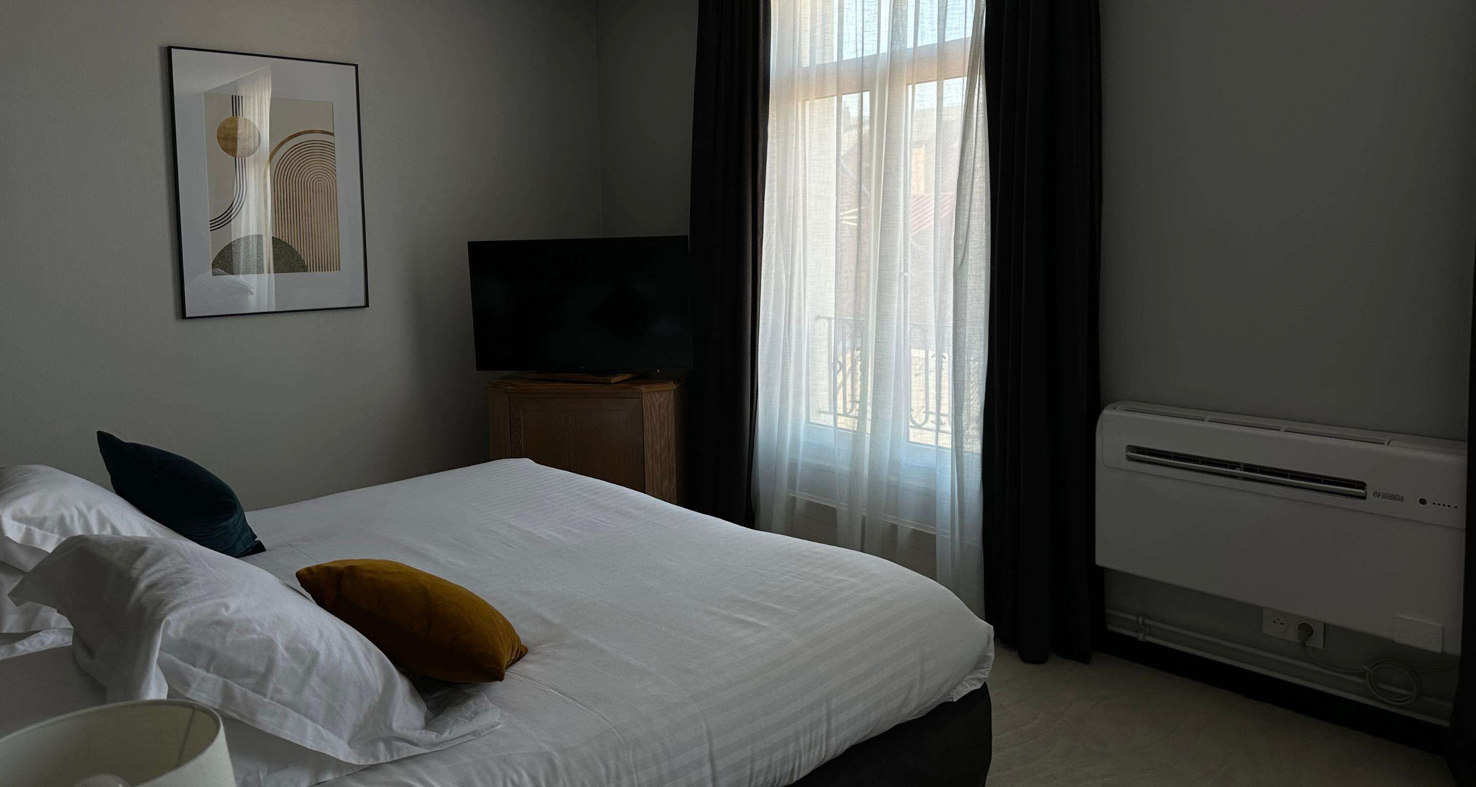 A hotel room with a bed, a flat-screen TV, a heater, and a window with curtains.