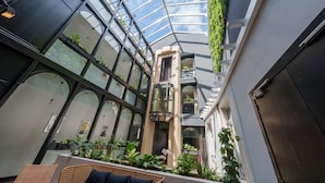 Een moderne atriumruimte met glazen dak, kamerplanten, rieten meubels en een centrale lift.
