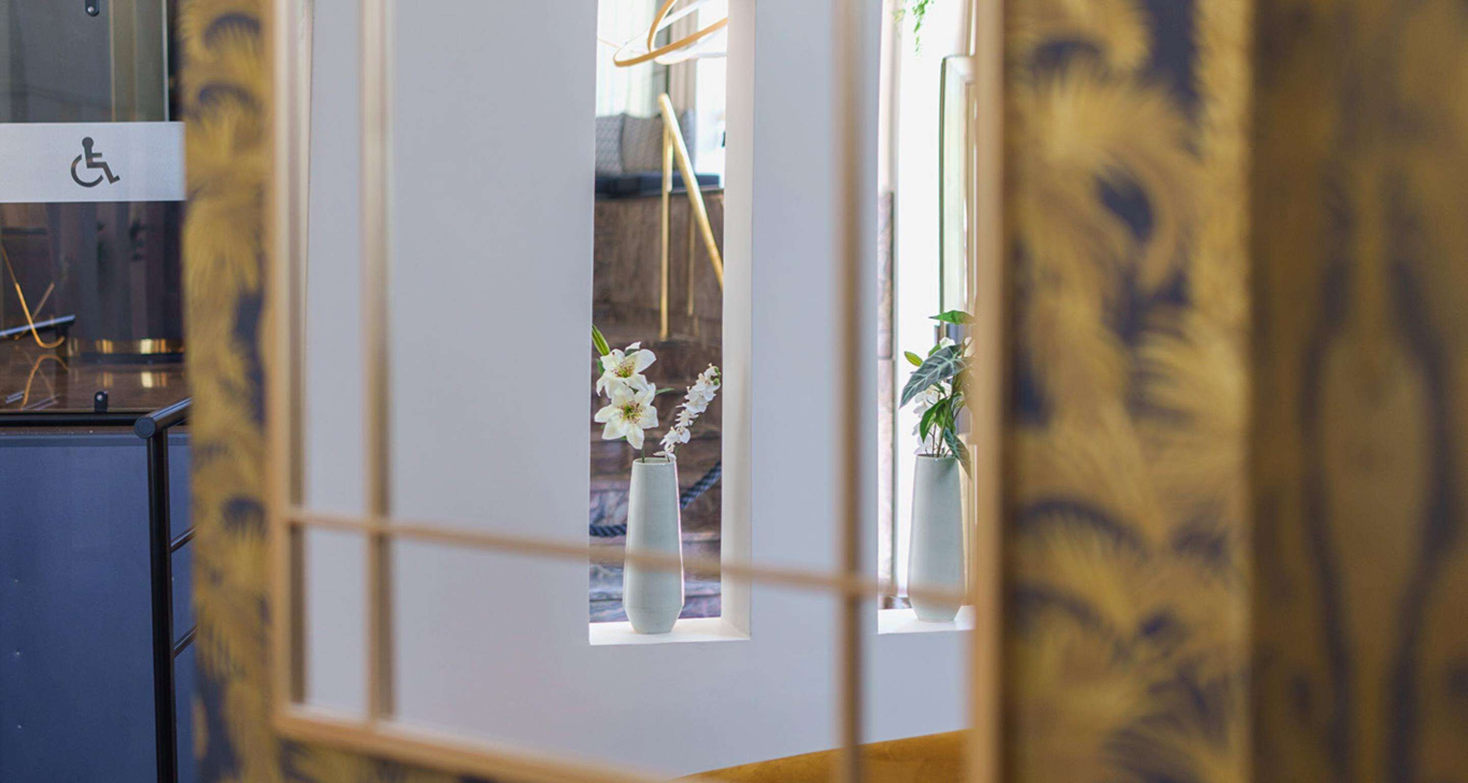 Ein Zimmer mit Rollstuhlgerechter Kennzeichnung, einem Fenster mit weißer Vase und Blumen sowie Blick auf eine Pflanze draußen.