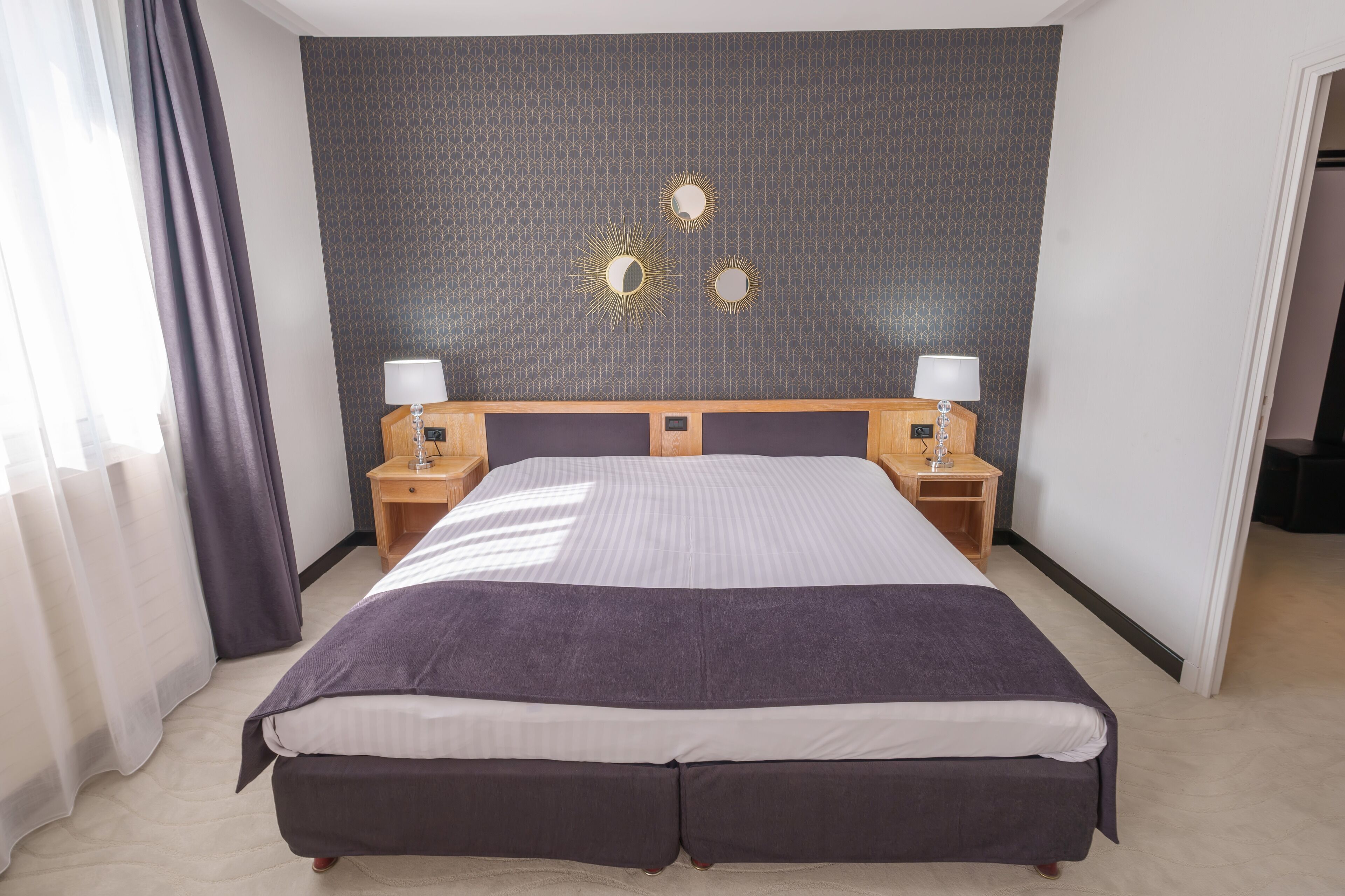 Habitación de hotel con una cama grande, dos mesitas de noche con lámparas, un cabecero oscuro con estampado y vistas al pasillo.