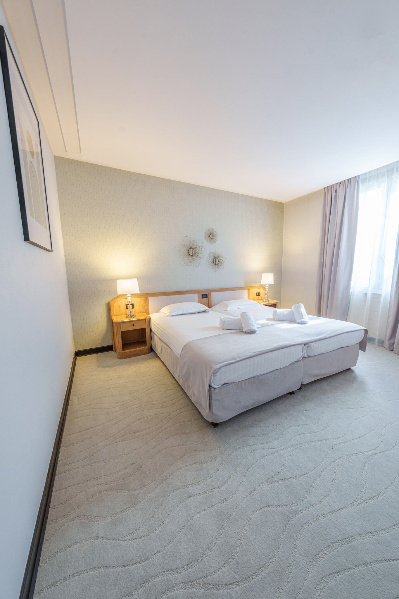 Habitación de hotel con una cama grande, dos mesitas de noche con lámparas, un adorno en la pared y una ventana con cortinas.