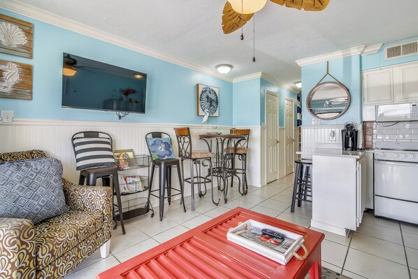 A living room with a sofa, chairs, a coffee table, and a kitchen area with a refrigerator and stove. - Coastal Blessings by Ryson Vacation Rentals (Galveston)