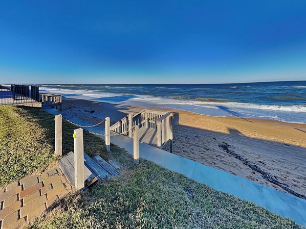 A beach with a wooden staircase leading down to the shore, a fence, and a clear view of the ocean.