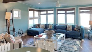 A living room with a blue sofa set, a white coffee table, and a view of the ocean through large windows. - Ocean Shore by Distinctive Beach Rentals (Flagler Beach)