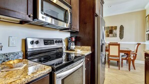 Una cocina con estufa de acero inoxidable, microondas y refrigerador, encimeras de granito, gabinetes de madera y un área de comedor con mesa y sillas.