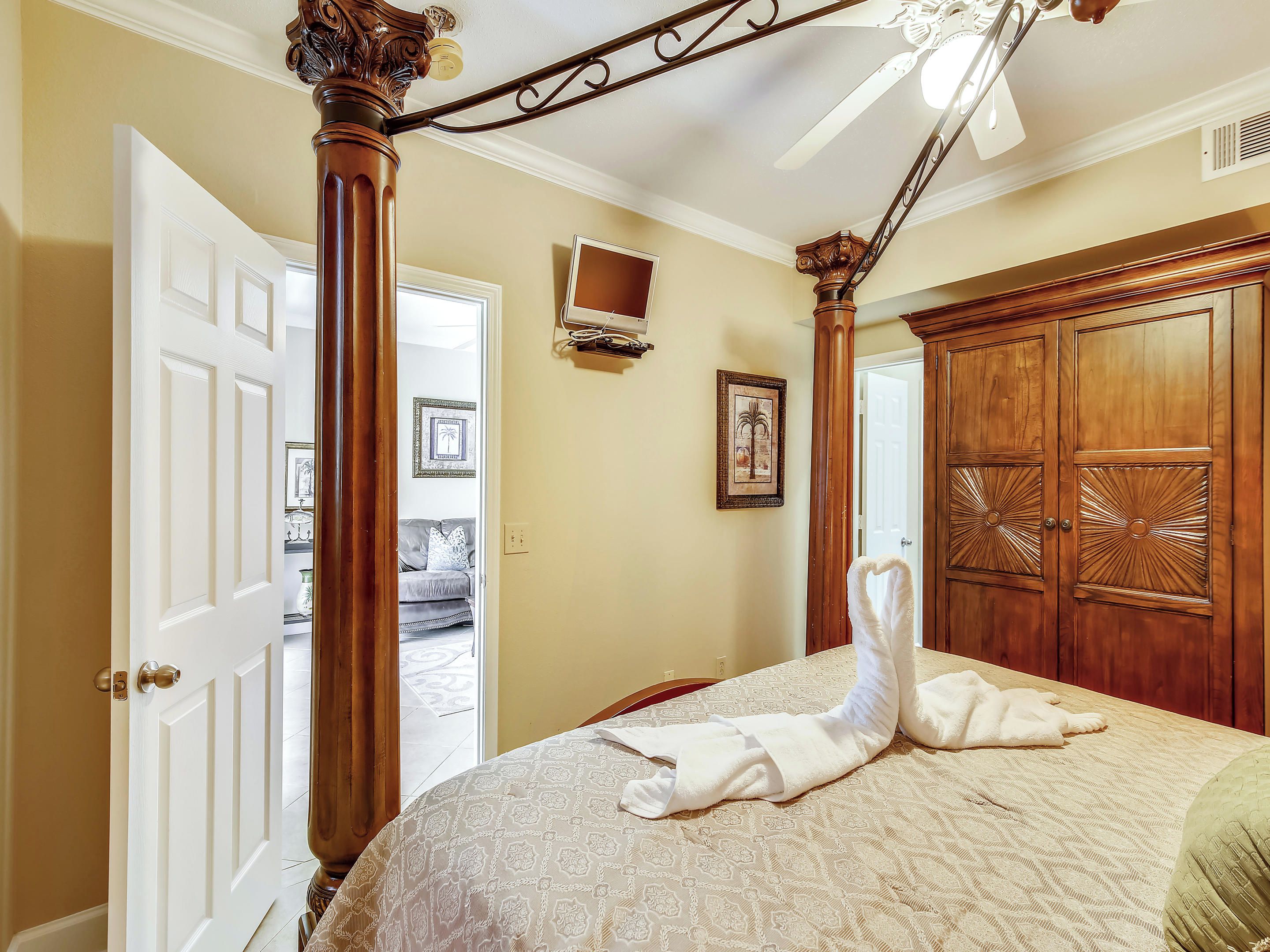 A bedroom with a bed, a wooden wardrobe, a ceiling fan, and a door leading to another room.
