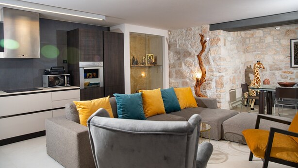 Ein modernes Wohnzimmer mit einem grauen Sofa, gelb-taubengrünen Kissen, einem Glastisch und einer Steinwand.