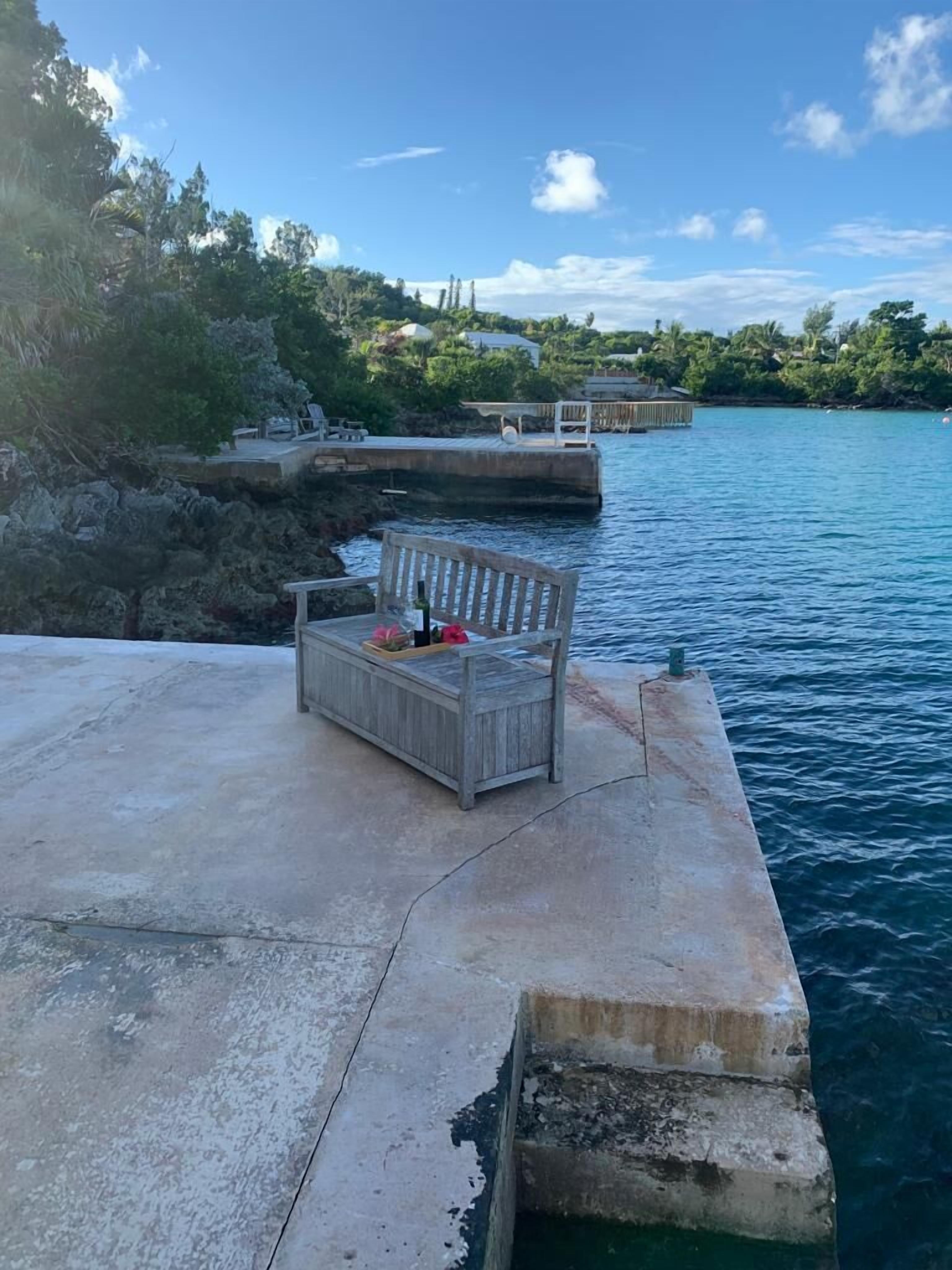 Une jetée en béton avec un banc en bois et une petite table, d’où l’on peut admirer une étendue d’eau calme et une rive lointaine parsemée de verdure et de bâtiments.