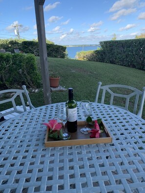 Mesa redonda con una botella de vino y dos copas sobre una bandeja, dispuesta al aire libre con vistas a un cuerpo de agua y follaje en el fondo.