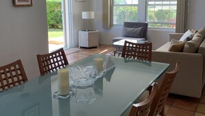 A dining area with a glass table, wooden chairs, and a small cabinet with a lamp.