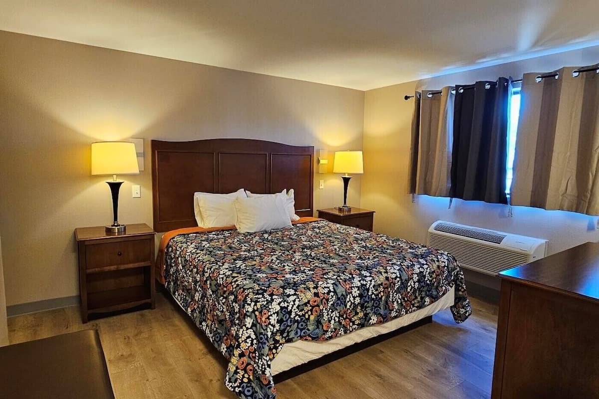 A hotel room with a bed, two bedside tables with lamps, a flat-screen TV, a window with curtains, and an air conditioning unit.