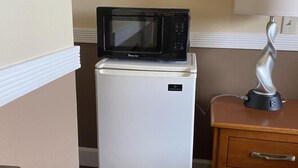 A compact kitchenette with a white refrigerator, a black microwave, and a wooden nightstand with a lamp. - Farwest Motel (Everett)
