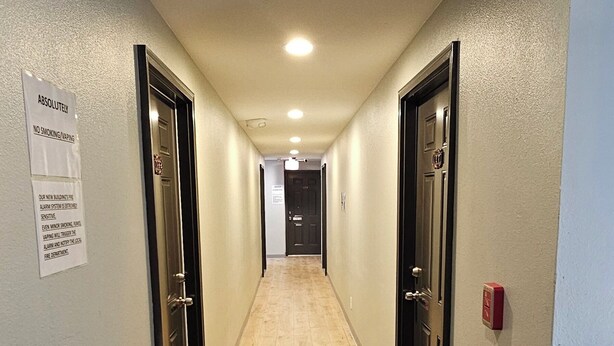 A hallway with wooden flooring, doors on both sides, a fire alarm on the ceiling, and a no-smoking sign on the wall. - Farwest Motel (Everett)