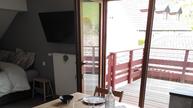 Habitación compacta con una mesa de comedor de madera, una silla, un taburete pequeño, un radiador, un televisor montado en la pared y una puerta corrediza de vidrio que da a un espacio exterior con barandilla roja.