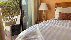 Une chambre d’hôtel avec un lit, une table de chevet avec une lampe, un fauteuil en osier et une vue sur les arbres à travers une porte vitrée.