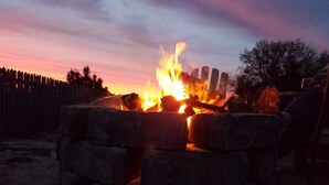 A campfire with logs stacked in a circular arrangement, surrounded by a wooden fence and trees in the background.