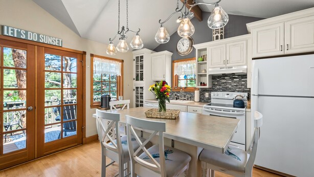 A modern kitchen with white cabinets, a central island with bar stools, pendant lighting, and a view through large glass doors. - Cozy cabin with Hot tub Fireplace Fire pit Full kitchen Close to town and RMNP. (Estes Park)