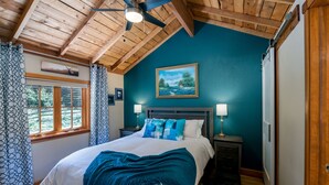 A bedroom with a bed, two bedside tables with lamps, a ceiling fan, a window with curtains, and a painting on the wall. - Cozy cabin with Hot tub Fireplace Fire pit Full kitchen Close to town and RMNP. (Estes Park)