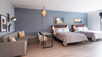 Une chambre d’hôtel moderne avec un canapé gris, une petite table, deux lits et des œuvres d’art abstraites accrochées au mur.