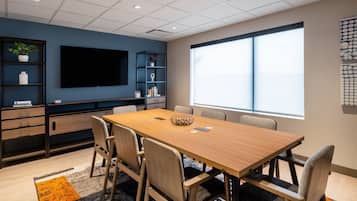 A conference room with a long table, chairs, a TV, shelves with plants, and a large window.