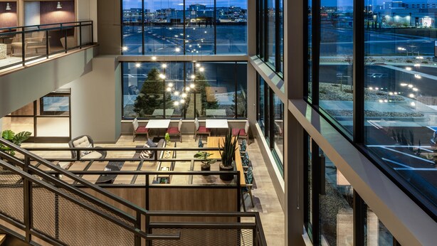 A modern hotel lobby with a high ceiling, large windows offering a view of the cityscape, and a seating area with tables and chairs.