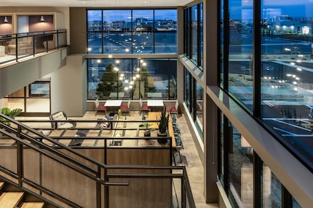 Un moderno vestíbulo de hotel con techos altos, amplias ventanas que ofrecen vistas panorámicas de la ciudad y un área de descanso con mesas y sillas.