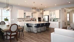 A modern kitchen with white cabinets, a central island with bar stools, pendant lights, and a dining area with a wooden table and chairs.