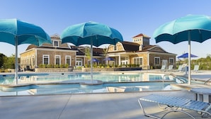 A swimming pool area with multiple umbrellas, lounge chairs, and a building with a brick facade in the background.