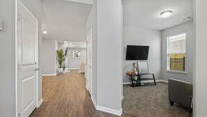 A hallway with a wooden floor, a white door, a grey sofa, a TV mounted on the wall, and a window with blinds. - New Modern Home, Lakefront, Park & Pool, spacious 4br/3ba very safe place<br> (Baton Rouge)