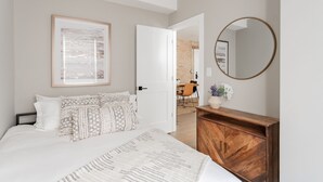 Une chambre à coucher comprenant un lit, une commode en bois, un miroir rond et une porte donnant sur une salle de bain.