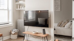 Un salon moderne avec un téléviseur à écran plat fixé au mur, une table basse en bois sur laquelle se trouvent un bol et un livre, un radiateur, un panier blanc contenant des vêtements pliés, et un lit avec des coussins à motifs en arrière-plan.